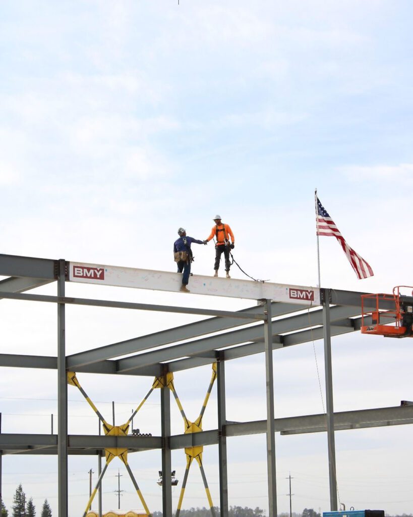 BMY-construction-worker-giving-a-hand-to-another-worker-atop-some-scaffolding-with-the-American-flag-in-the-background